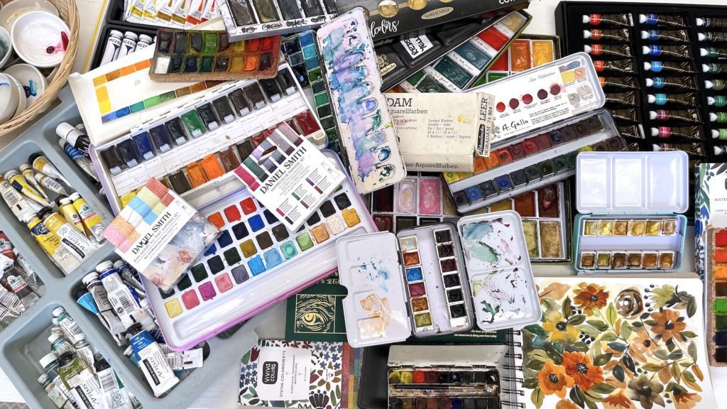 Too many watercolor supplies on a cluttered desk
